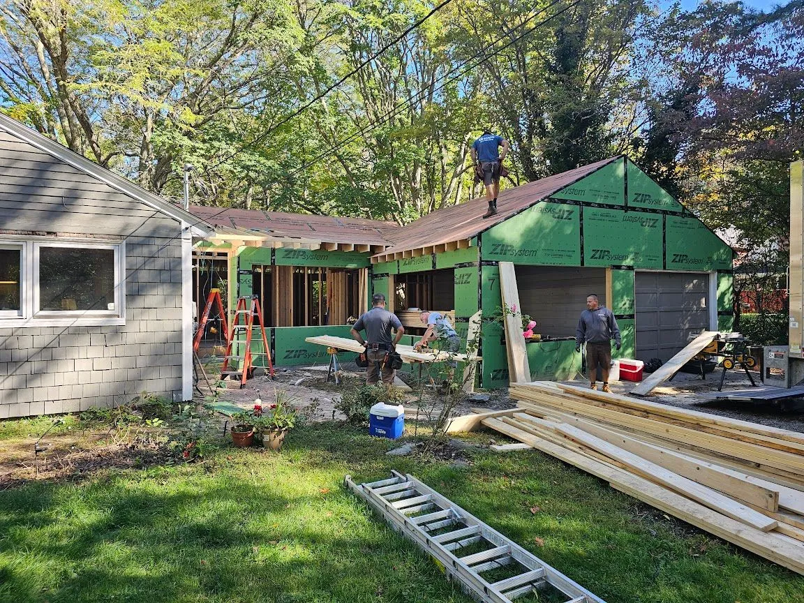 Addition framing tied into an existing gray house, with a crew working on the roof and lumber on the lawn