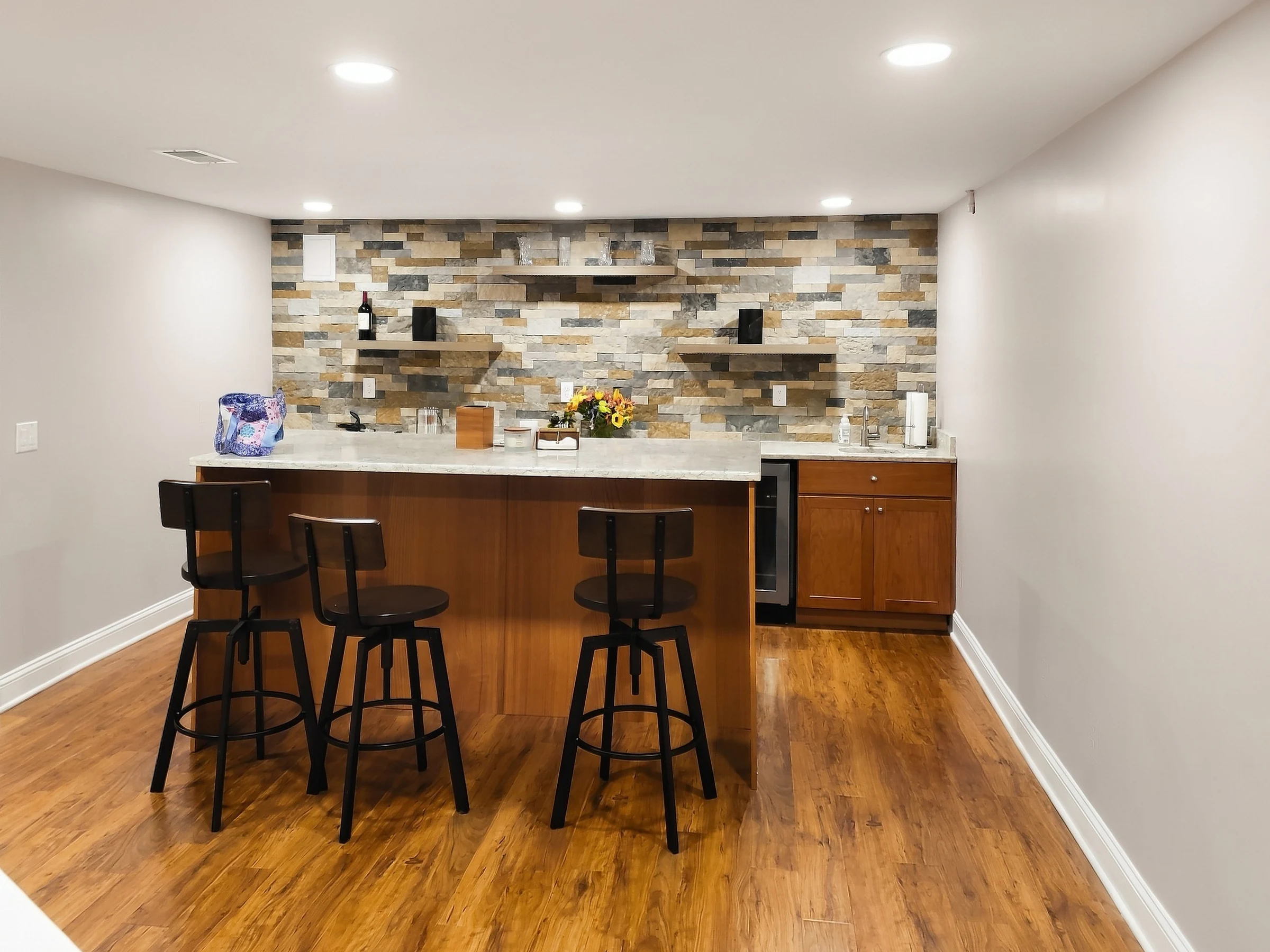 Basement wet bar with stools, wood cabinets and a patterned backsplash.