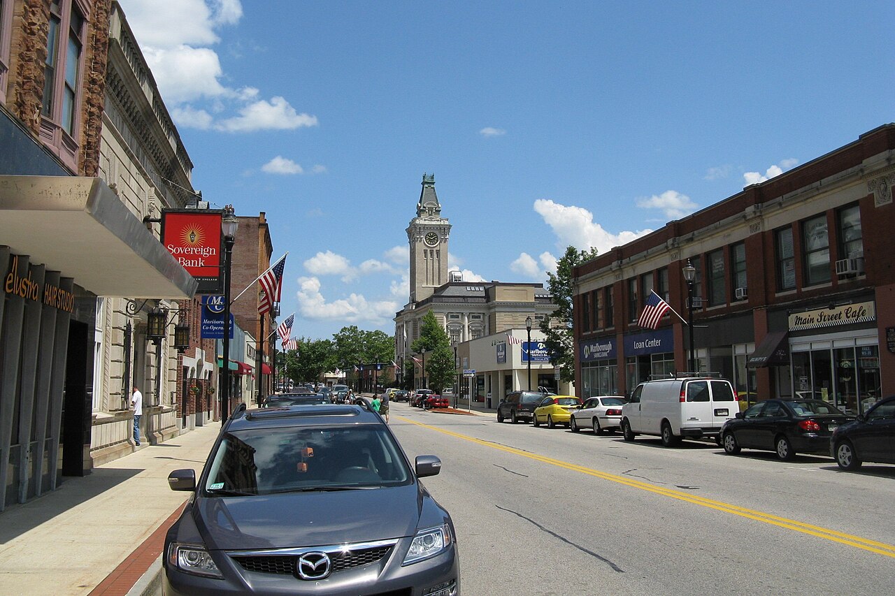 Main Street in Marlborough looking east, with brick storefront blocks, parked cars and a clock tower in the distance